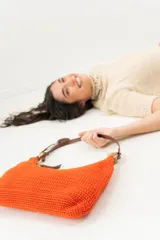 Cartera tipo shoulder bag tejida a mano con punto crochet en raffia color naranja. Tiene asa de cuero marrón y cierre superior con cremallera.