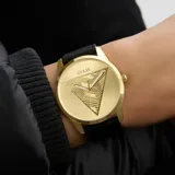 Reloj Guess con caja dorada y esfera dorada con relieve de triángulo texturizado. Posee agujas blancas y correa de silicona negra.