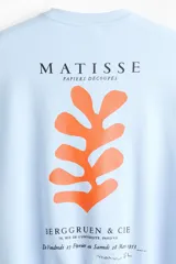 Buzo celeste de corte holgado con hombros caídos y cuello redondo. Presenta un pequeño estampado en el pecho con la inscripción "Matisse" y un detalle gráfico en color naranja.