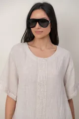 Vestido midi de lino color beige, con mangas cortas, cuello redondo, alforzas verticales en el pecho y detalle de volados en el ruedo.