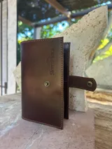 Sobre de cuero color marrón oscuro, con costuras visibles en contraste y cierre a presión metálico. Presenta el logo grabado "SierraMora" en la parte superior frontal.