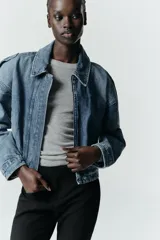 Cazadora de denim estilo crop con cuello solapa, manga larga y cierre frontal con cremallera y botón metálico. Presenta bolsillos de vivo en el delantero y detalle de trabillas en los hombros.