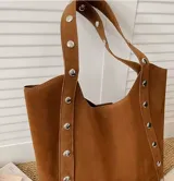 Cartera tipo tote de símil gamuza color camel con diseño amplio, correas de hombro integradas y detalles de tachas metálicas en los laterales.
