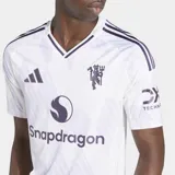 Camiseta de fútbol Adidas del Manchester United, modelo segunda equipación 25/26, para hombre. Diseño blanco con cuello redondo y detalles en violeta. Presenta un patrón sutil en la tela, logo de Adidas, escudo del club con acabado metalizado y sponsor Snapdragon en el frente.