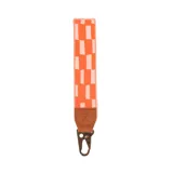 Llavero de muñeca con cinta de nylon estampada con diseño geométrico en tonos rosados y naranja, detalle de cuero sintético marrón con logo grabado y gancho metálico.