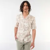 Camisa de manga corta color beige con estampado abstracto de hojas blancas, cuello camisero abierto y cierre frontal de botones.
