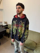 Hoodie oversize de color negro con estampado gráfico en pecho y mangas en tonos amarillos y verdes. Presenta un forro interior de capucha en color rojo con textura tejida y logo bordado en la manga.