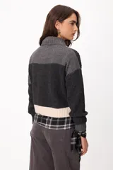 Sweater de punto con diseño patchwork en tonos gris, grafito y crudo, con cuello alto tejido. Presenta una estructura de bloques de color y texturas variadas, incluyendo detalles de trenzas y tejido acanalado.