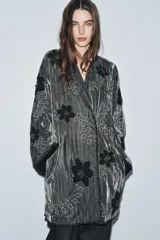 Kimono de terciopelo gris con acabado texturizado, cuello redondo y manga larga. Presenta bordados florales en hilo negro y metalizado, bolsillos delanteros y cierre frontal.
