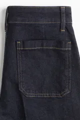 Pantalón de jean de tiro alto, color azul oscuro (índigo), con pierna ancha y corte cropped. Presenta bolsillos de parche grandes en la parte delantera y costuras en color ocre.
