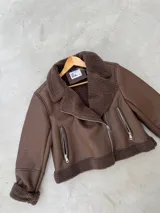 Campera tipo gamulán de cuero sintético en color marrón chocolate, con interior y solapas de corderito sintético al tono. Presenta cierre frontal cruzado y bolsillos laterales con cierre metálico.