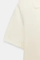 Top de punto color crudo, con cuello polo y escote en V. Cuenta con mangas cortas y detalle de puntilla con forma de onda en el cuello y los puños.