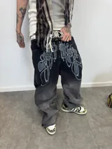 Jean de corte amplio con efecto de lavado degradado de negro a gris. Presenta un estampado gráfico estilo tribal en la parte superior delantera y cierres laterales metálicos a lo largo de las piernas que permiten ajustar la amplitud del corte. Incluye bolsillos amplios y botones personalizados.