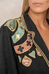 Blazer negro de corte recto con solapas decoradas con ribete dorado trenzado. Presenta bordados artesanales coloridos en el hombro derecho y detalles de flecos con cuentas turquesas en los bolsillos frontales. Cuenta con cierre de un solo botón.