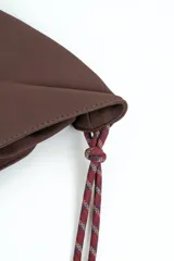 Bolso tipo morral color marrón de diseño curvo, con manija de cuerda trenzada en tonos burdeos y detalles de nudos en los extremos.