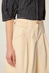 Bermuda de tiro alto en color crudo, confeccionada en lona de algodón. Presenta un corte acampanado con pliegues en la cintura, bolsillos delanteros inclinados y costuras decorativas en contraste color camel.