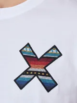 Remera blanca de algodón orgánico con cuello redondo y manga corta. Presenta un estampado en el pecho con una cruz formada por dos tablas de surf en tonos rojo, azul y negro.