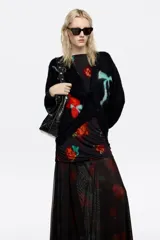 Vestido midi de corte ajustado con falda superpuesta de tul, escote barco y mangas largas tipo murciélago. Presenta un diseño estampado con motivos florales en tonos rojos y verdes sobre fondo negro, con detalles fruncidos en la cintura.