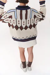 Campera tipo bomber tejida con diseño jacquard geométrico en tonos azul marino, blanco, rojo y verde. Presenta cierre frontal completo, cuello, puños y ruedo acanalados en color verde y blanco, con corte oversized y hombros caídos.