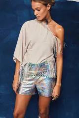 Top beige asimétrico de un solo hombro con mangas murciélago amplias, adornado con detalles de flecos delicados en el escote y en el ruedo inferior.