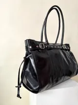 Bolso grande de cuero marrón con textura tipo cocodrilo, estructura rígida y asas para llevar al hombro. Tiene una tira con tachas y hebillas en cada extremo, también lleva lazos en los costados.