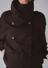 Chaqueta marrón de corte recto y diseño crop, confeccionada en mezcla de algodón y poliamida con forro interior de poliéster. Presenta cuello alto tipo embudo, doble botonadura frontal, manga larga y dos bolsillos laterales.
