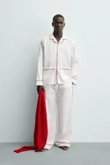 Pijama de dos piezas confeccionado en popelín de algodón blanco, con vivos de ribete en color bordó. Incluye una camisa de manga larga con cuello bowling, bolsillos de plastrón y cierre frontal de botones, y un pantalón de corte relajado con cintura elástica y bolsillos laterales.