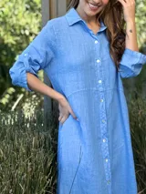 Vestido camisero largo de lino blanco, con cuello camisero, manga corta enrollada y cierre frontal de botones de nácar. Presenta aberturas laterales en el bajo.