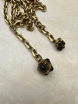 Collar de cadena de eslabones gruesos en tono dorado, con diseño tipo lazo que termina en dos dijes colgantes: uno esférico con detalles labrados y piedras negras, y otro con forma de flor decorado con cristales negros.