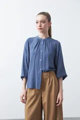 Camisa de color celeste claro con silueta relajada y mangas largas. Presenta un cuello redondo con pequeña abertura frontal y botones, además de un delicado trabajo de tablas en los hombros que aporta textura al diseño.