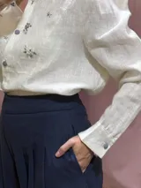 Camisa de lino 100% natural, de color blanco, con mangas largas abullonadas y bordados artesanales a mano en el frente. Cuenta con cuello mao y cierre frontal de botones grises.
