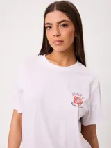 Remera blanca de corte holgado, con cuello redondo y mangas cortas. Presenta un pequeño estampado en el pecho con diseño floral y tipografía.
