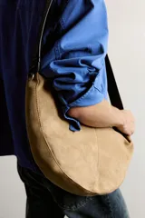 Bolso de hombro de gamuza marrón con diseño curvado, compartimento principal con cierre de cremallera y asa larga de cuero negro.