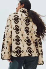 Buzo oversized de sherpa suave en color beige con estampado geométrico en tonos marrones y detalles en tejido revestido. Presenta cuello elevado, medio cierre frontal y bolsillos laterales.