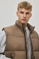 Chaleco acolchado color tostado, de nylon guateado, repelente al agua y rompevientos. Cuenta con cuello elevado, cierre bidireccional con protección para el mentón, bolsillos laterales con cierre y un bolsillo interior con botón a presión. El bajo es ajustable con cordón elástico y topes.