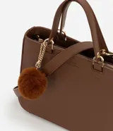 Cartera estilo satchel de tamaño mediano, confeccionada en poliuretano color marrón con textura tipo cuero. Cuenta con asas de mano reforzadas, correa larga desmontable, cierre superior de cremallera y un colgante decorativo de pompón de pelo sintético.
