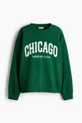 Buzo de cuello redondo en color verde, con diseño gráfico estampado en blanco en la parte frontal. Presenta hombros caídos, corte holgado y terminaciones acanaladas en cuello, puños y ruedo.