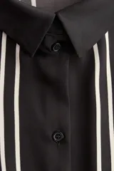 Camisa de corte oversized en tejido negro con leve brillo y diseño de líneas geométricas en color blanco. Cuenta con cuello clásico, cierre frontal de botones y mangas largas anchas.