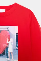 Camiseta de algodón color rojo, corte regular fit, cuello redondo y manga corta. Presenta una estampa fotográfica en el centro con dos personas sosteniendo un gran corazón rojo inflable. Incluye texto con nombre y copyright.