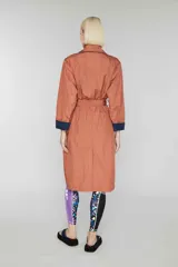 Trench largo cruzado color ladrillo, sin forro, con cinturón reversible azul marino y detalles de color de contraste ocultos. Tiene botones en el frente, bolsillos de vivo oblicuos y capucha oculta.