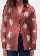 Camisa de manga larga en color terracota con diseño semitransparente. Presenta cuello camisero, cierre frontal con botones y bordados florales en color blanco distribuidos por toda la prenda.