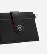 Billetera compacta Satinato de poliuretano color negro, con solapa frontal con botón a presión, detalles en rojo y argolla lateral metálica.