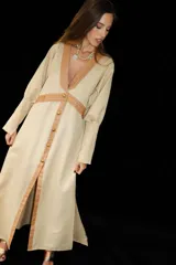 Vestido largo color beige, con escote en V profundo y solapas en tono marrón claro. Presenta una hilera de botones dorados en el frente y detalles de tiras horizontales en el mismo tono marrón claro en la cintura.