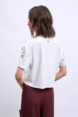 Remera blanca de algodón con cuello redondo y mangas cortas. Presenta apliques metálicos y aberturas circulares en los hombros y el frente.