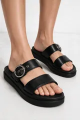 Sandalias planas de dos tiras anchas en color negro, con suela track y hebilla metálica circular plateada en la tira superior.