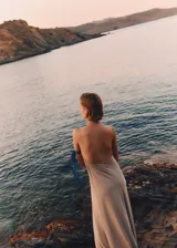 Vestido largo de corte evasé en color beige, con cuello halter cruzado y espalda abierta. Presenta un diseño minimalista y fluido con cierre de botones en la parte posterior del cuello.