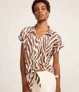 Blusa de manga corta confeccionada en viscosa, con cuello camisero, cierre frontal con botones y detalle de lazo para anudar en el bajo. Presenta un estampado híbrido entre follaje y animal print de cebra en tonos marrón y blanco.