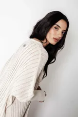 Poncho de punto color crudo con calce suelto y relajado. Confeccionado en tejido con lúrex que aporta un sutil brillo, presenta cuello en V, mangas anchas y textura acanalada.