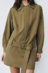 Vestido corto verde militar de cuello solapa y manga larga, con bolsillos de plastrón en el delantero y cierre frontal con cremallera oculta por sopata. Cuenta con un cinturón incorporado del mismo tejido con hebilla metálica y aberturas laterales en el bajo.