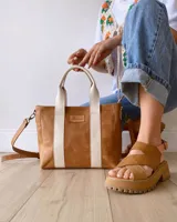 Cartera tipo tote de cuero color camel con detalles de bandas verticales en color crema. Cuenta con doble asa de mano, correa larga ajustable y extraíble, y cierre superior. Incluye etiqueta de cuero con logo grabado en el frente.